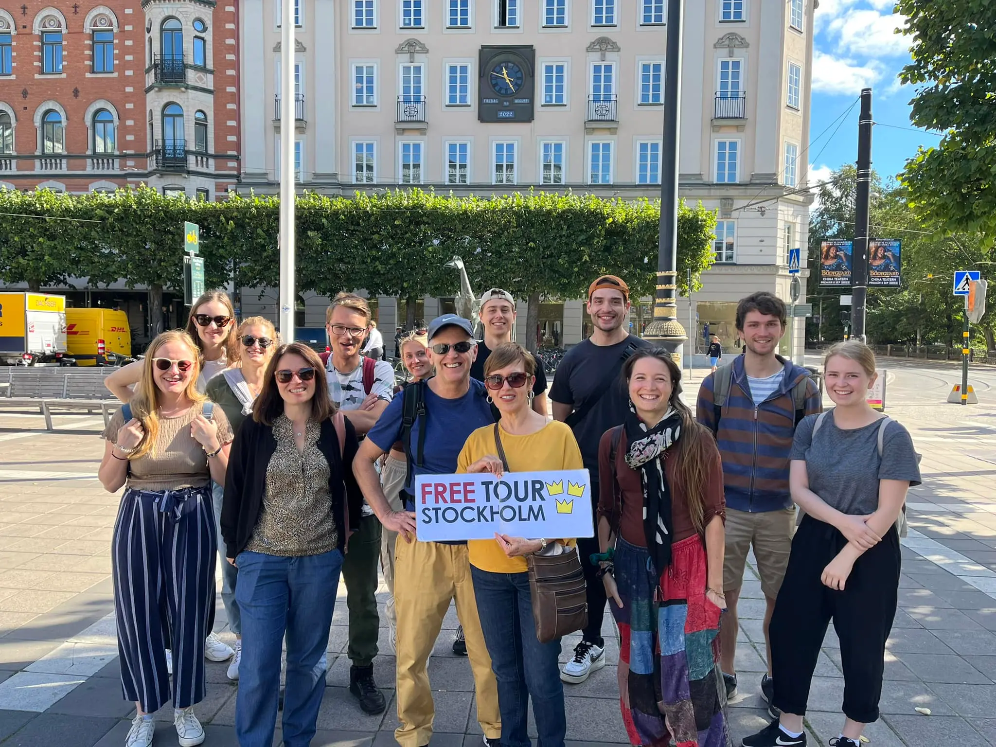 A tour group is posing at a sunny Norrmalmstorg square in Stockholm, and a young woman is holding a sign that says Free Tour Stockholm.